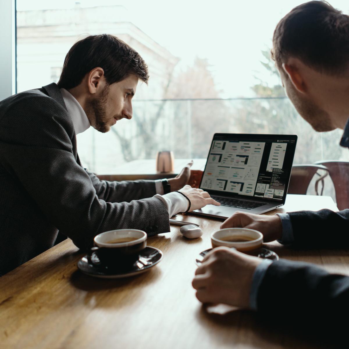 Two businessmen talking about new opportunities sitting with laptop at desk, planning project, considering business offer, sharing ideas while drinking coffee together